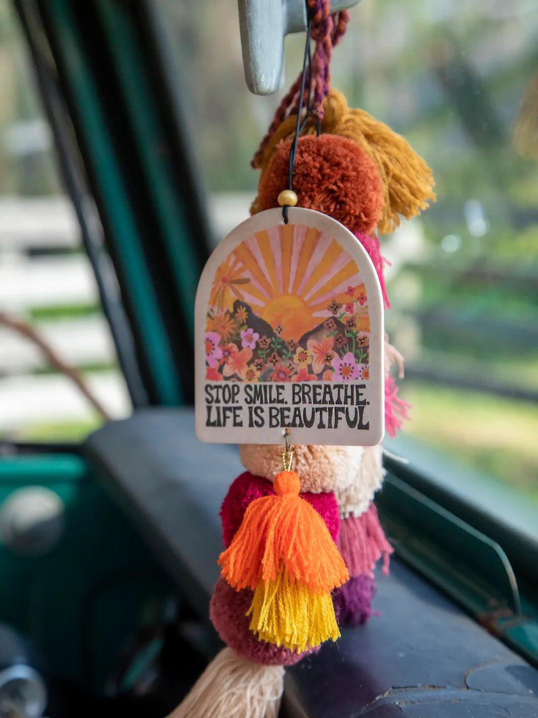 Natural life strawberry-scented essential oil car air freshener that reads "Stop. Smile. Breathe. Life Is Beautiful." It features double-sided artwork with sunset and scenery graphics and hanging tassels.