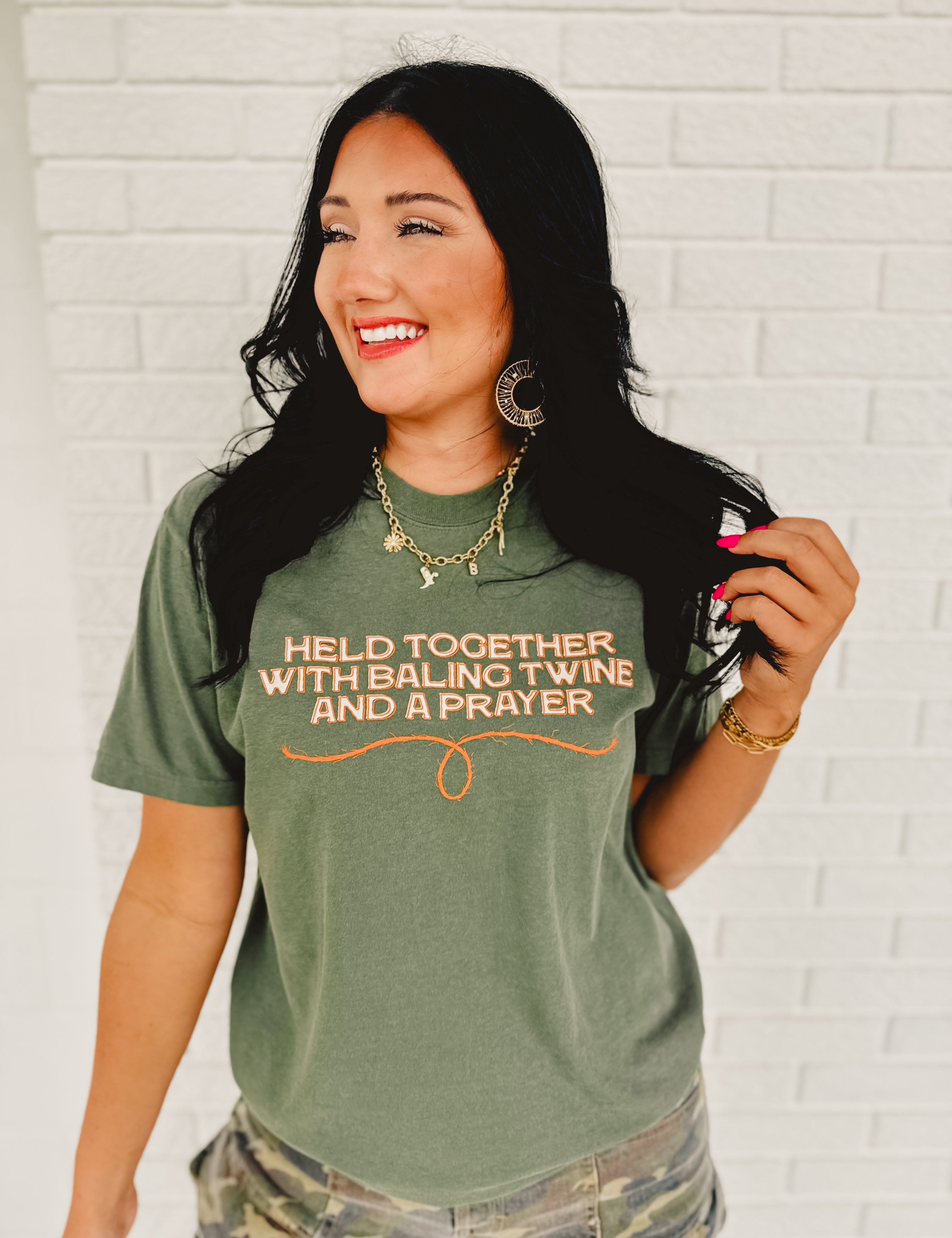 Moss green short sleeved rounded neck graphic tee that reads " Held Together With Baling Twine And A Prayer."