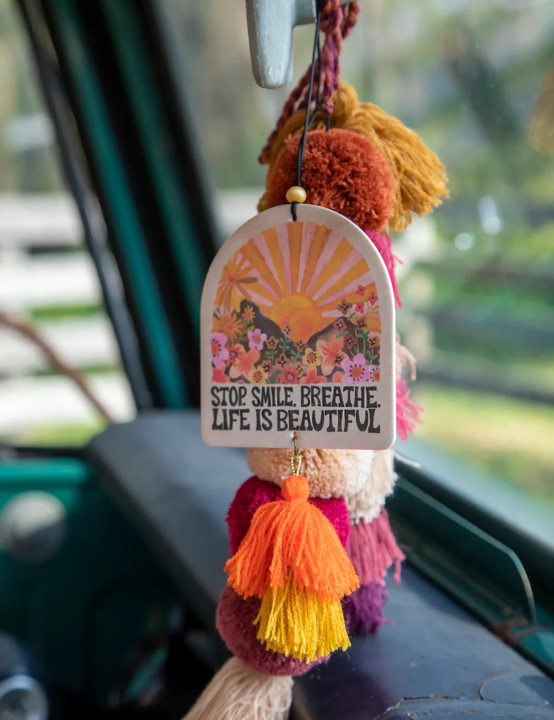 Natural life strawberry-scented essential oil car air freshener that reads "Stop. Smile. Breathe. Life Is Beautiful." It features double-sided artwork with sunset and scenery graphics and hanging tassels.